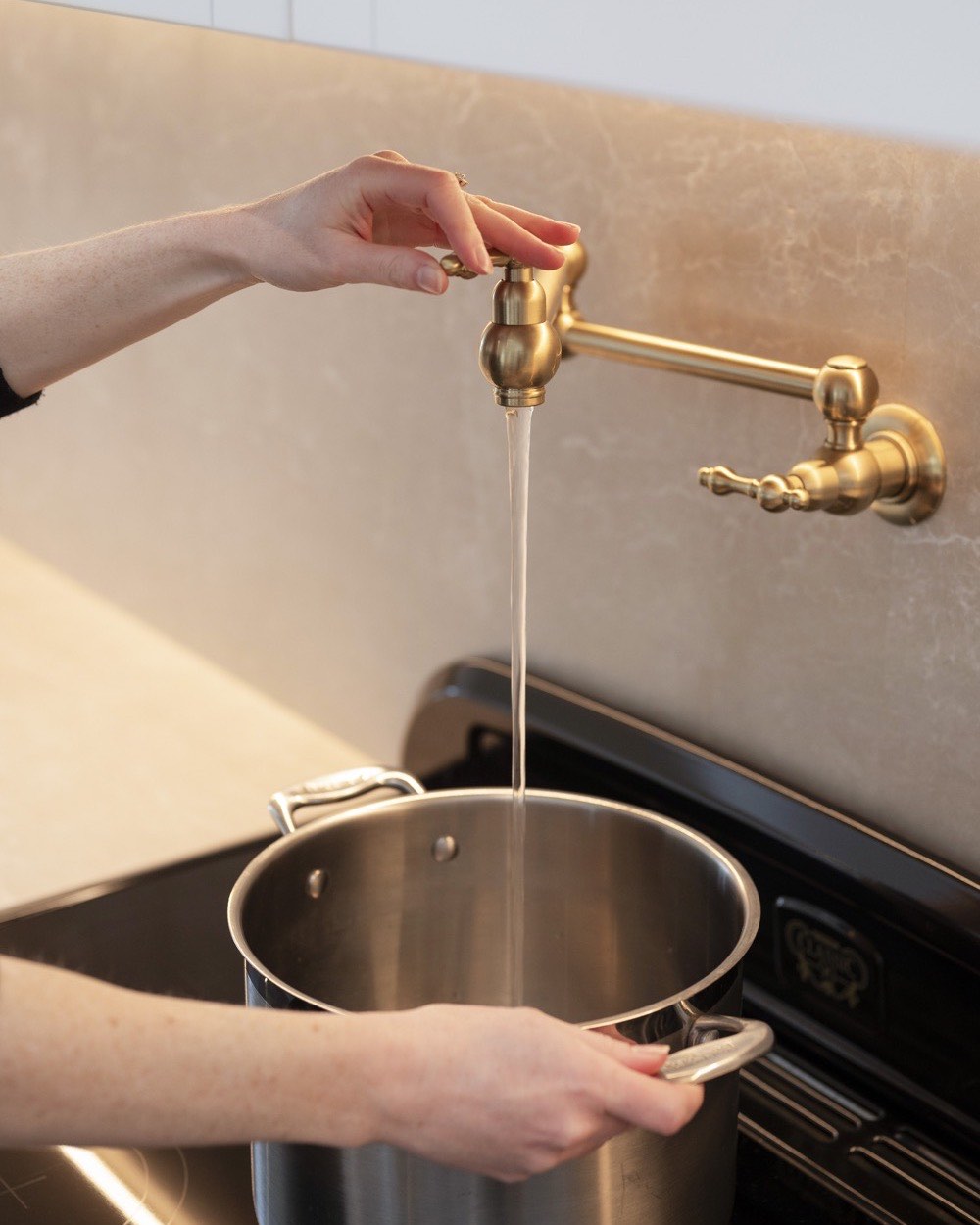 A hand filling a pot at a brass pot filler over a stone splashback